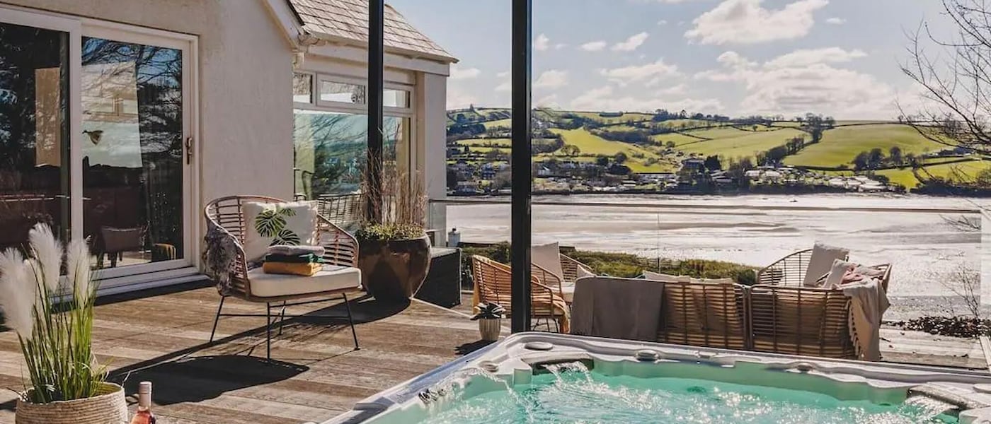 A waterfront view from the private deck of a white bungalow, where a hot tub bubbles in the foreground and a garden dining set overlooks the estuary.
