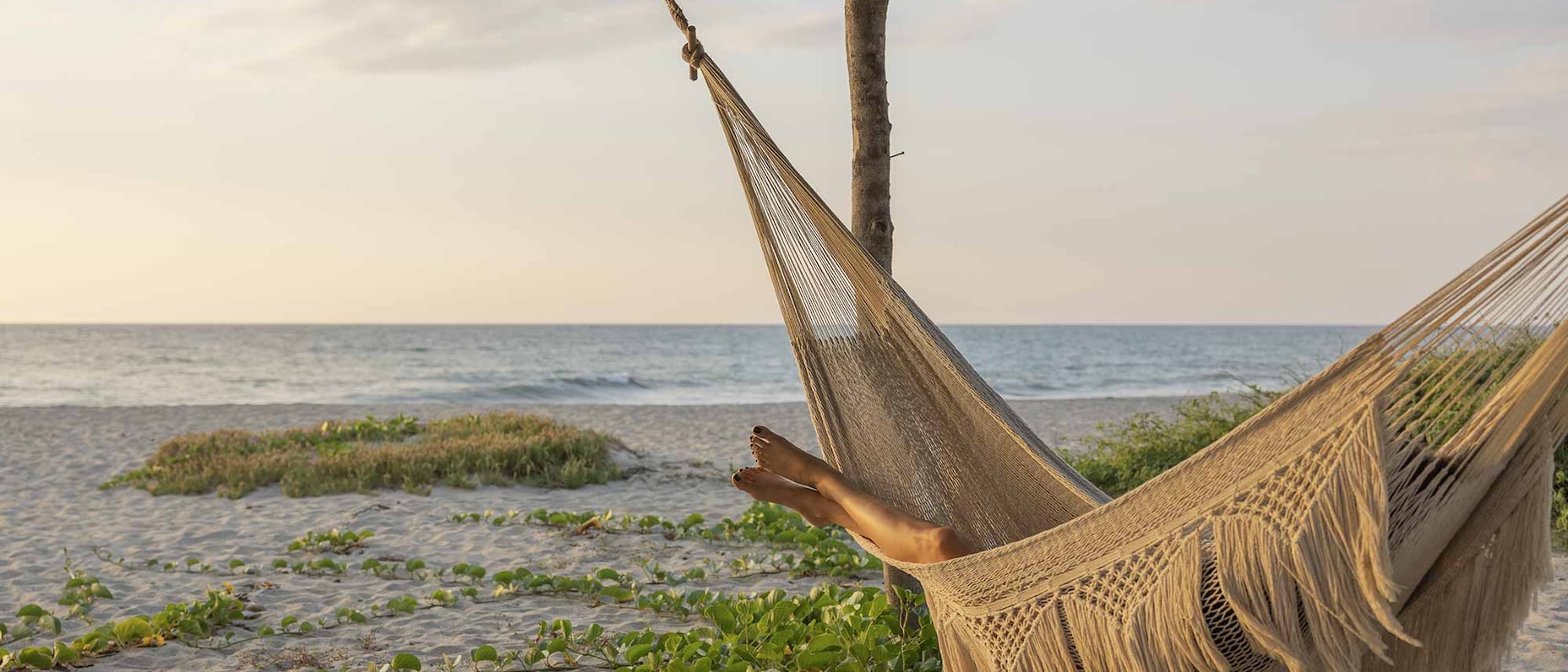 Una persona relajándose en una hamaca con flecos frente al mar en una playa tranquila. 