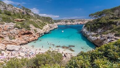 Udsigt over en solbeskinnet bugt, hvor folk soler sig på stranden og bader i det klare, blå hav. 