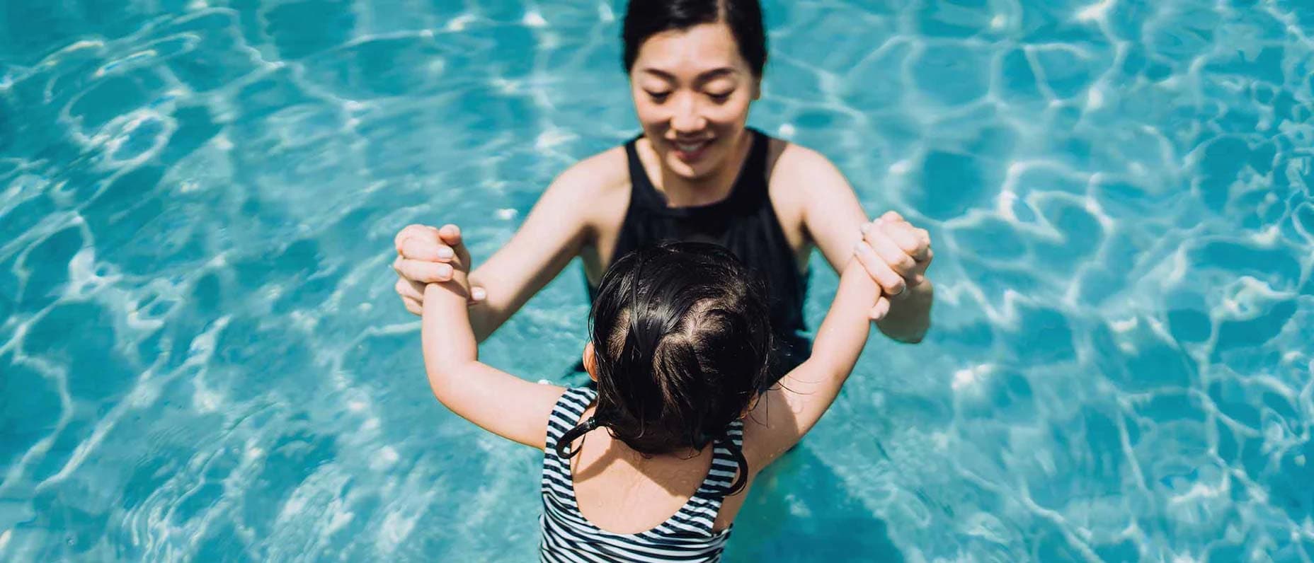 A woman standing waist-high in a swimming pool smiles as she holds hands with a little girl, seen from behind, who is sitting on the edge