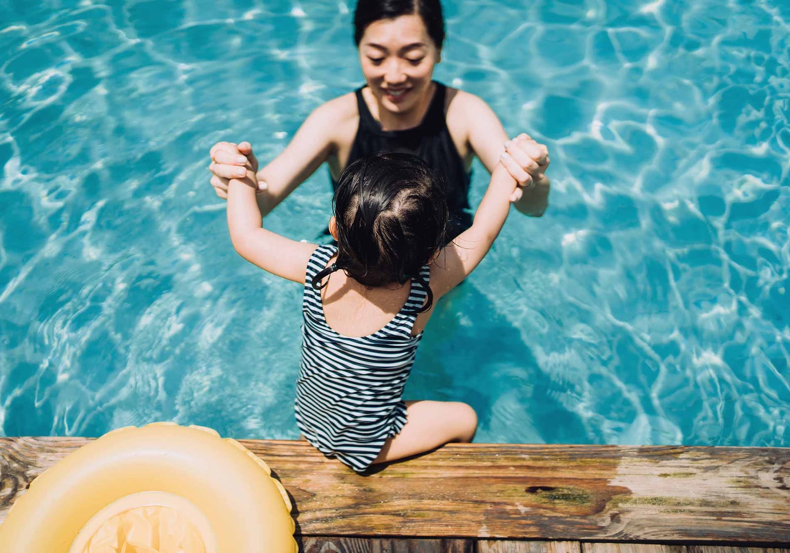 A woman standing waist-high in a swimming pool smiles as she holds hands with a little girl, seen from behind, who is sitting on the edge