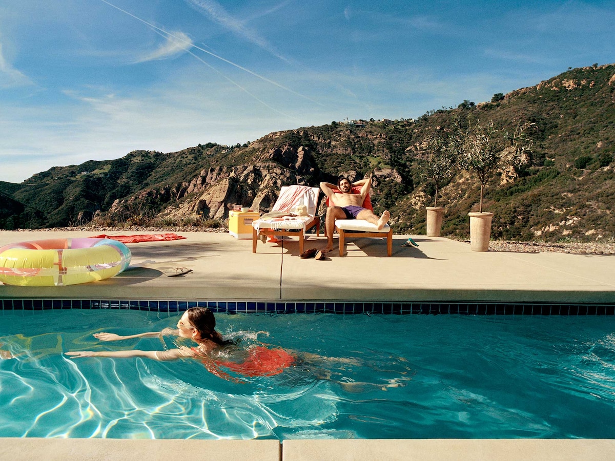 Une personne nage dans une piscine tandis qu’une autre se détend sur une chaise longue avec des montagnes en arrière-plan.