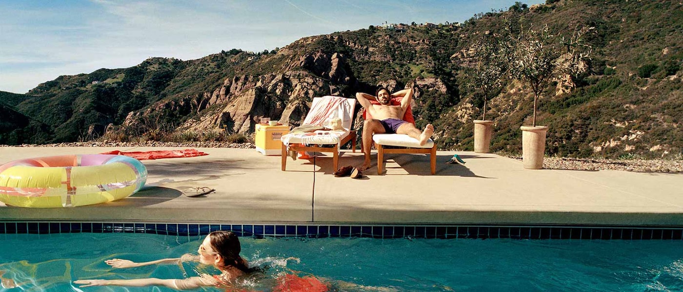 A woman swims in a pool while a man relaxes on a lounger with mountains in the background.