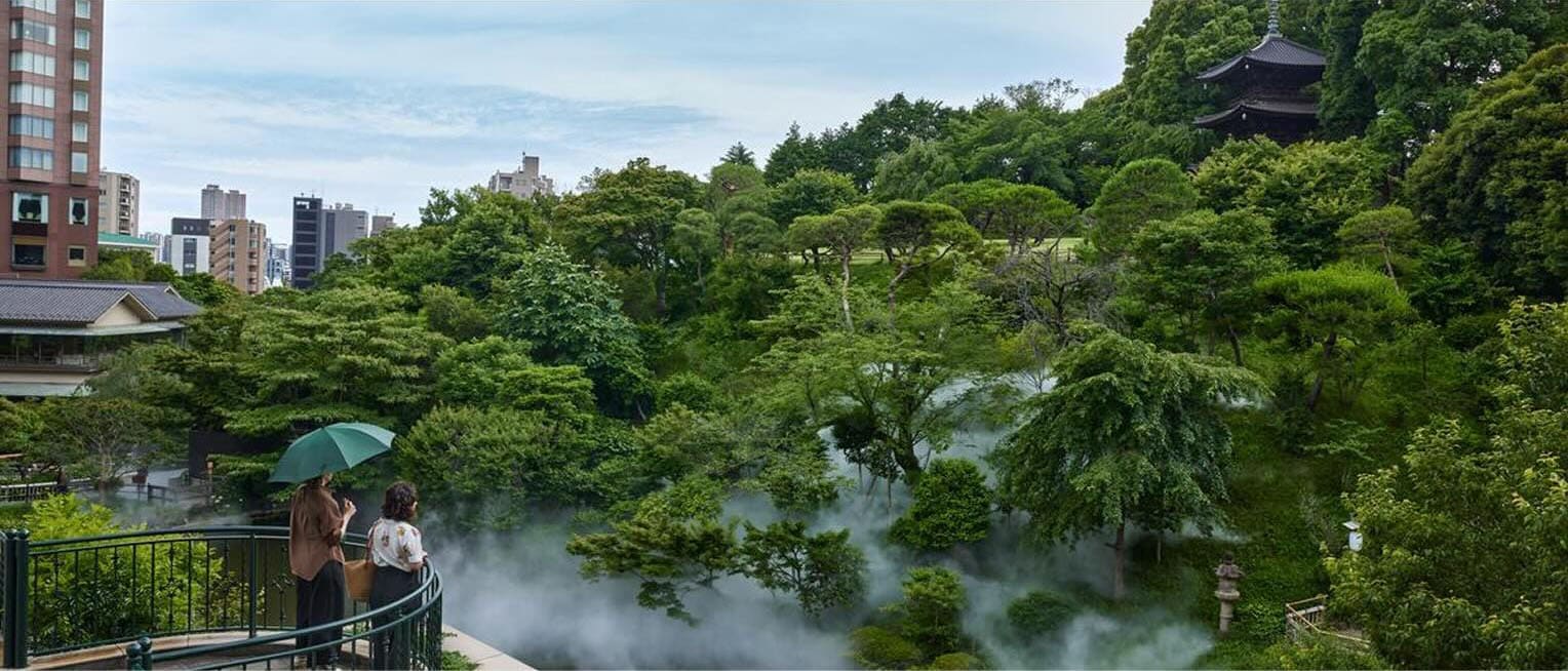 Deux personnes sont debout sur un balcon surplombant les jardins de l’Hotel Chinzanso Tokyo, avec des nuages entre les arbres, les bâtiments voisins en arrière-plan et le sommet de la pagode Entsukaku.