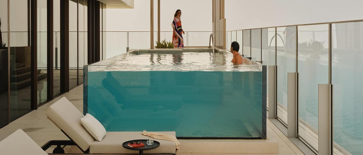 Des voyageurs profitent de la piscine aux parois de verre sur le balcon de l’Atlantis The Royal, avec au premier plan des effets personnels laissés sur des chaises longues.