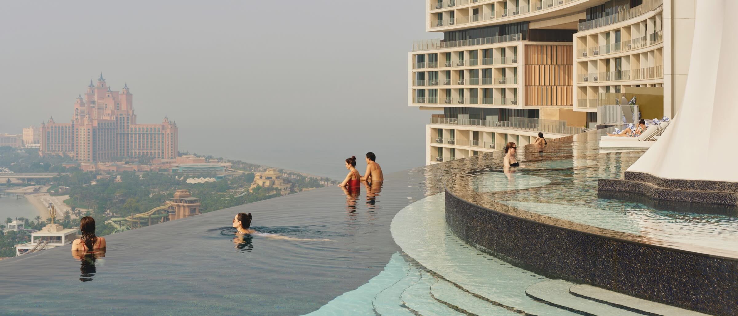Des voyageurs à l’Atlantis The Royal profitent de la piscine à débordement avec vue sur Dubaï. L’architecture moderne et incurvée de l’hôtel est également visible.