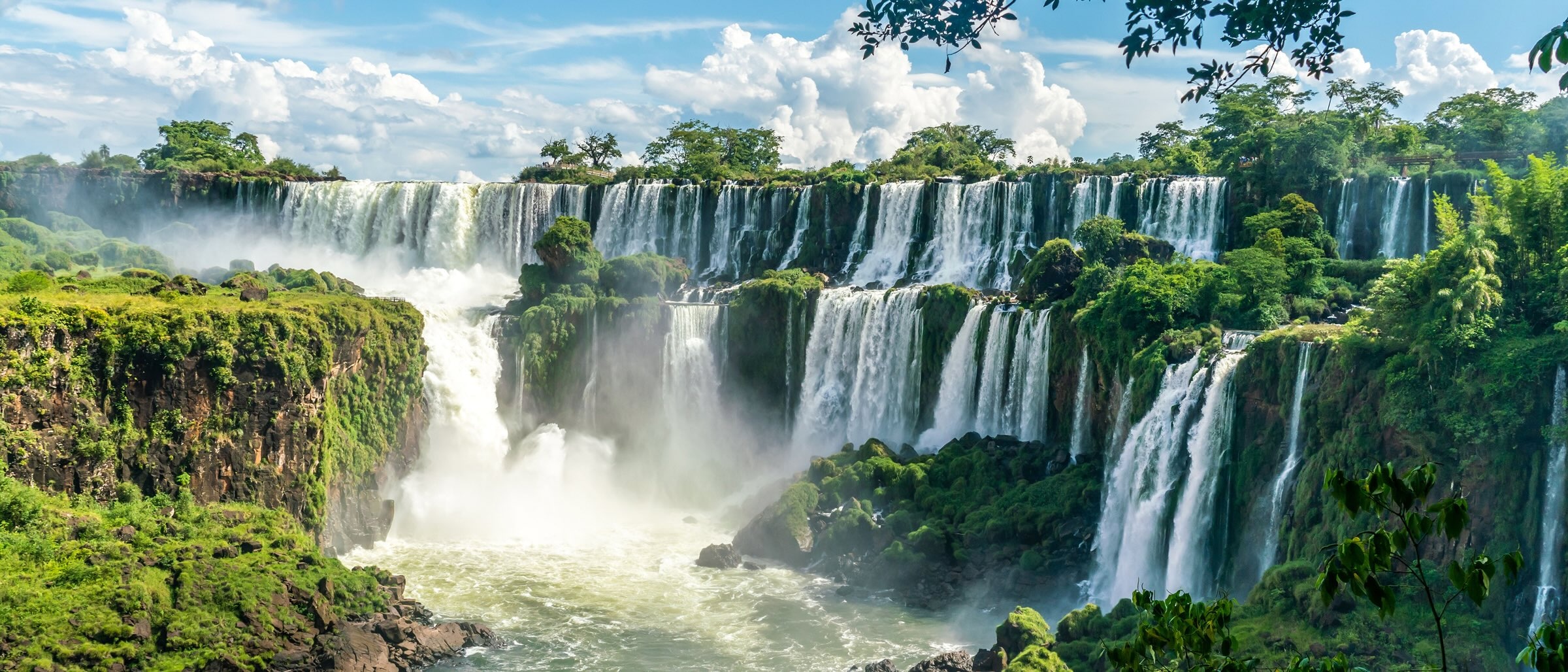 Panoramic view of Iguazu Falls cascading over lush green cliffs, surrounded by dense rainforest under a bright blue sky with scattered clouds.