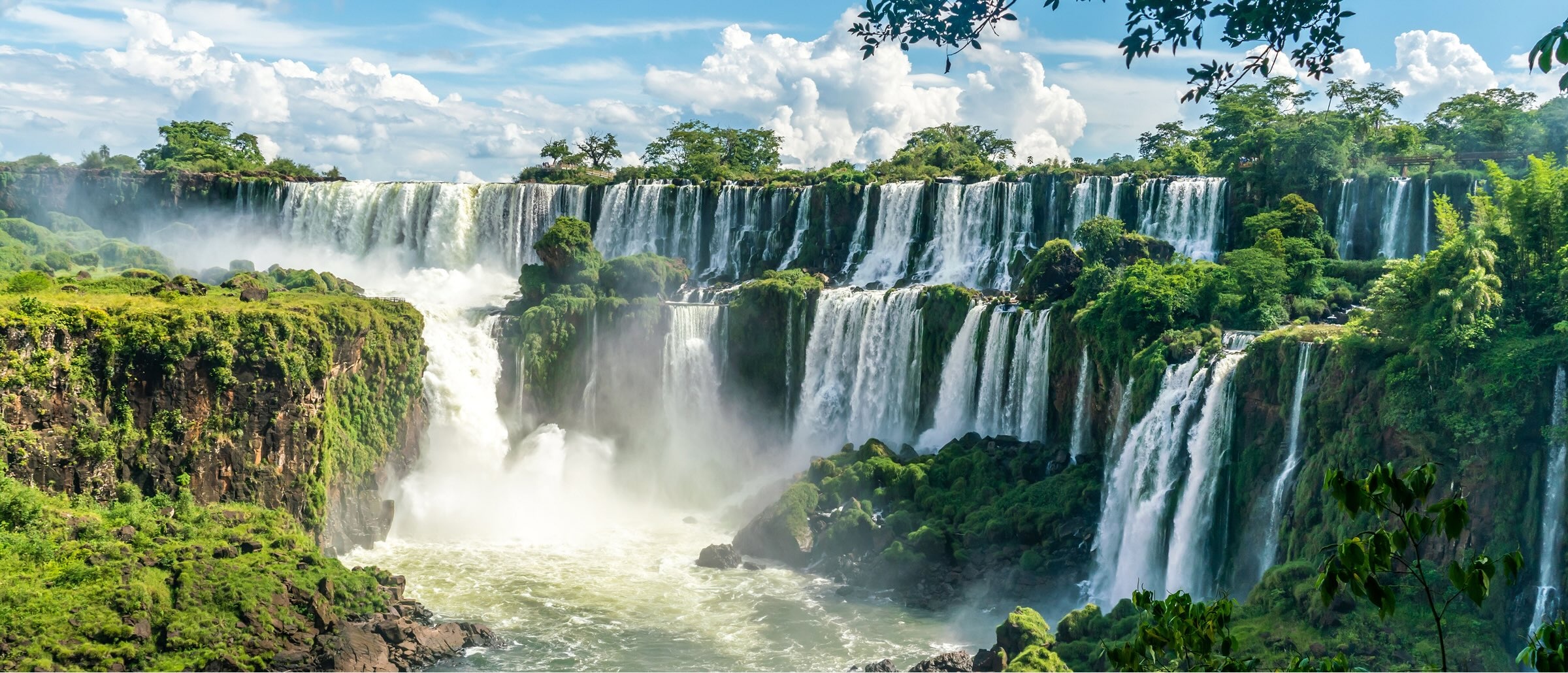 Panoramic view of Iguazu Falls cascading over lush green cliffs, surrounded by dense rainforest under a bright blue sky with scattered clouds.