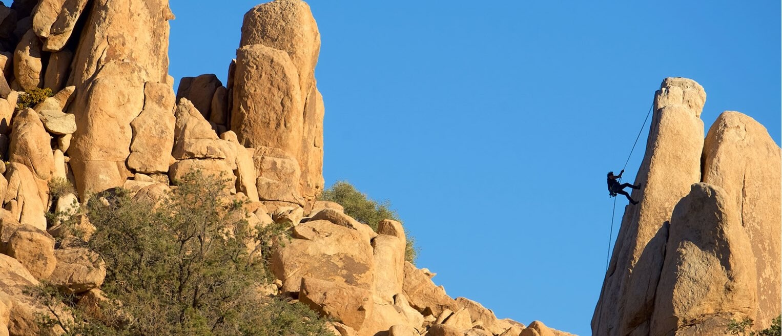 Silhouette of a rock climber scaling a steep sandstone spire under a bright blue sky in a desert terrain.