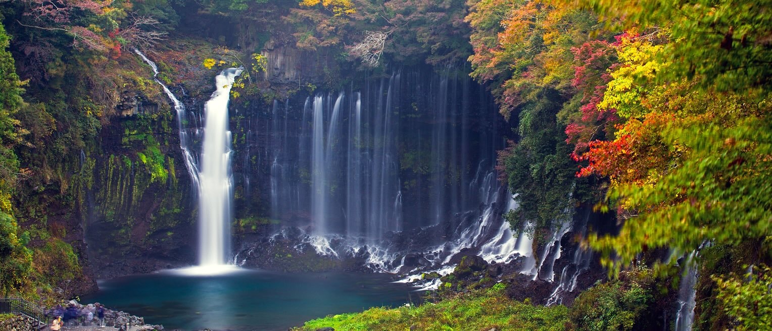 Delicate, curtain-like Shiraito Falls flowing into a calm pool, framed by vibrant autumn foliage in Japan.