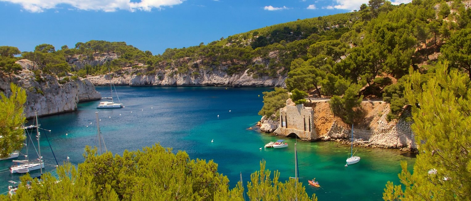 Turquoise inlet in the Calanques National Park, lined with pine trees, rocky cliffs and sail boats anchored in the bay.