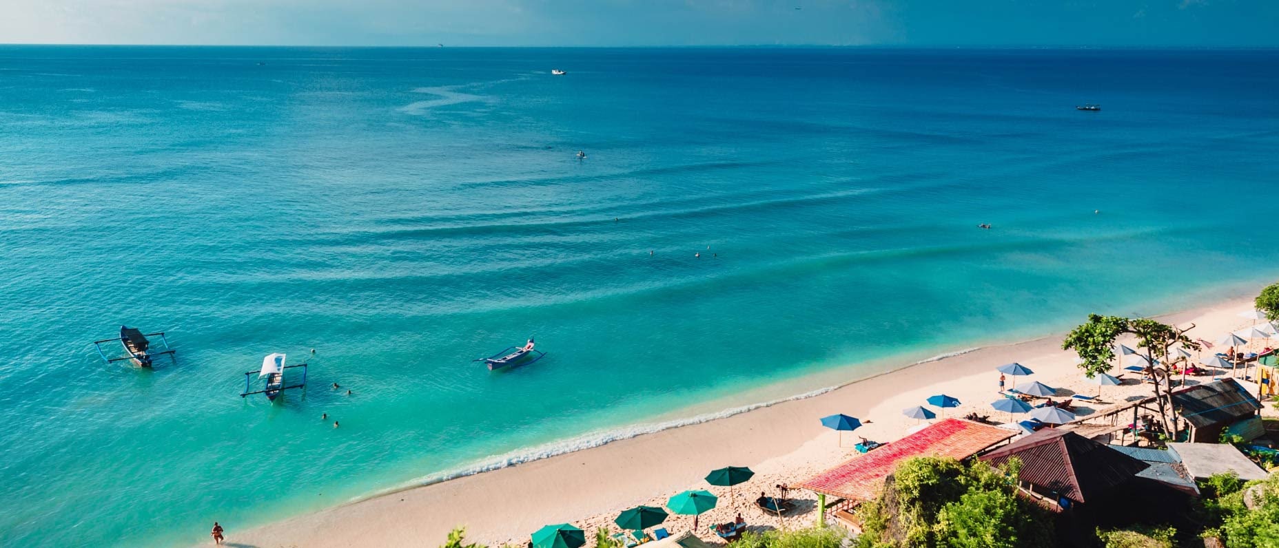 A vast expanse of turquoise sea laps gently onto a golden sandy beach. Umbrellas are scattered along the shoreline, and a boat floats on the calm water.
