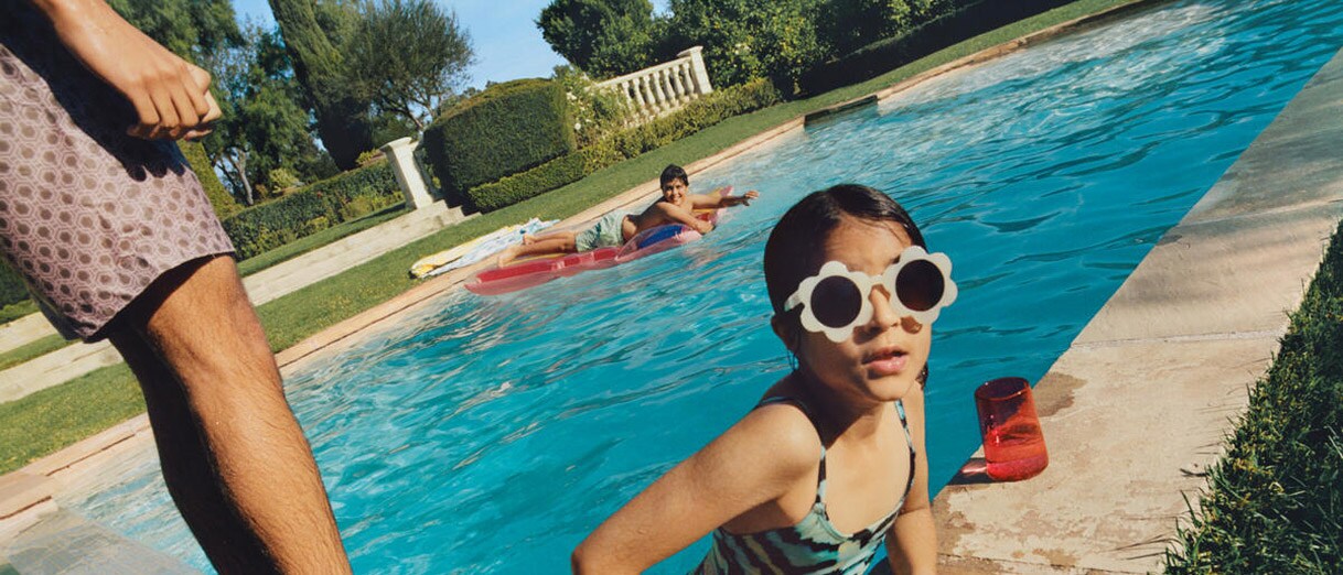 A boy on an inflatable and a girl in sunglasses enjoy a pool as an adult looks on