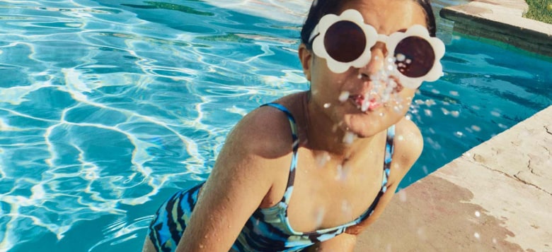A boy on a floatie and a girl in sunglasses enjoy a pool as an adult looks on