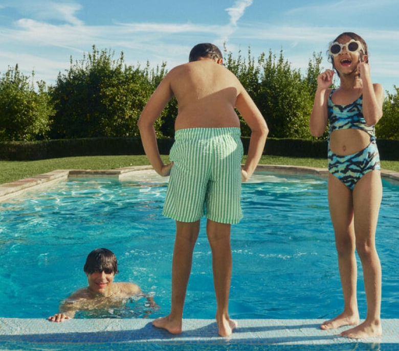 A girl in singlasses looks at the camera while a boy contemplates the water poolside