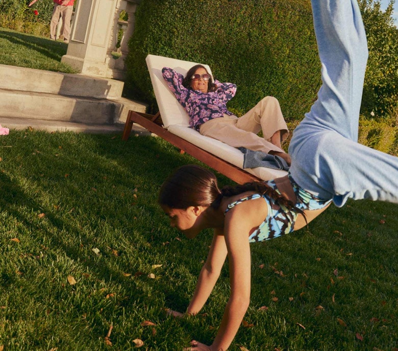 A girl does a cartwheel on a lush lawn as an adult reclines on patio furniture