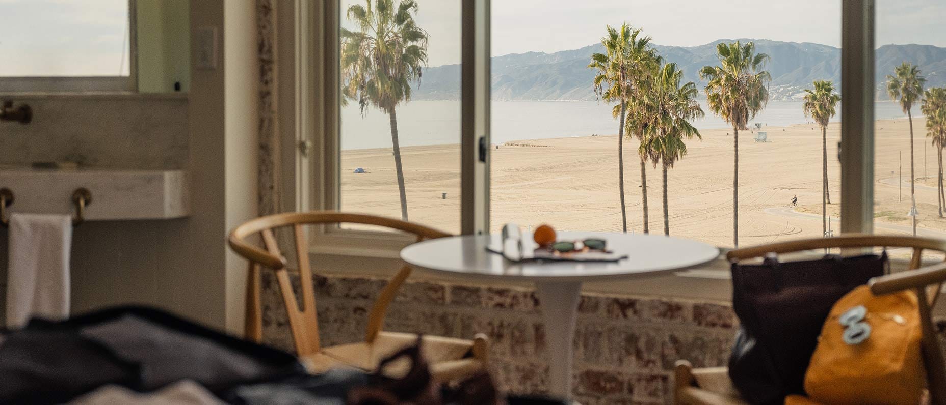 Vista de um quarto de hotel com paredes de tijolos e vista para uma praia tranquila cercada de palmeiras em um dia de sol. 