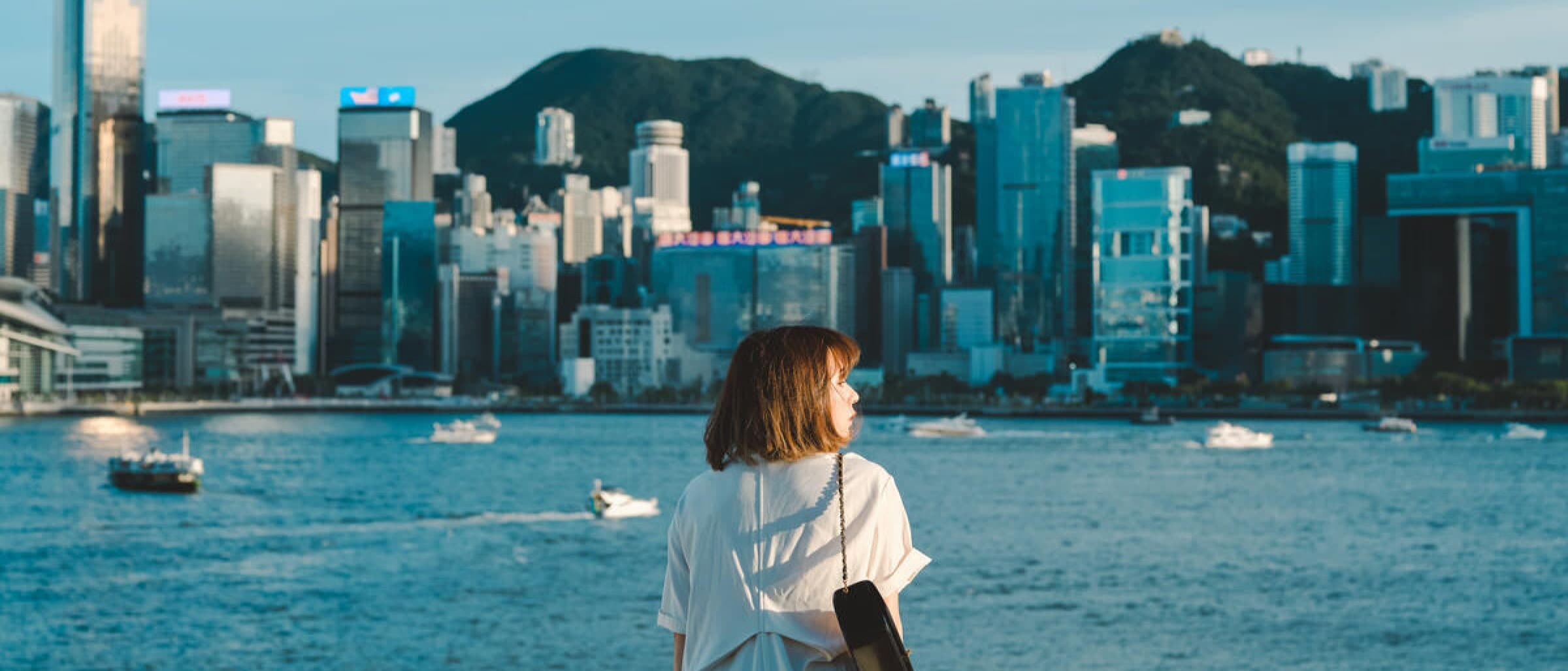 一個身穿白裙的女子站在海濱欄杆前,眺望維港對岸的香港天際線,背景有山,海中有船。