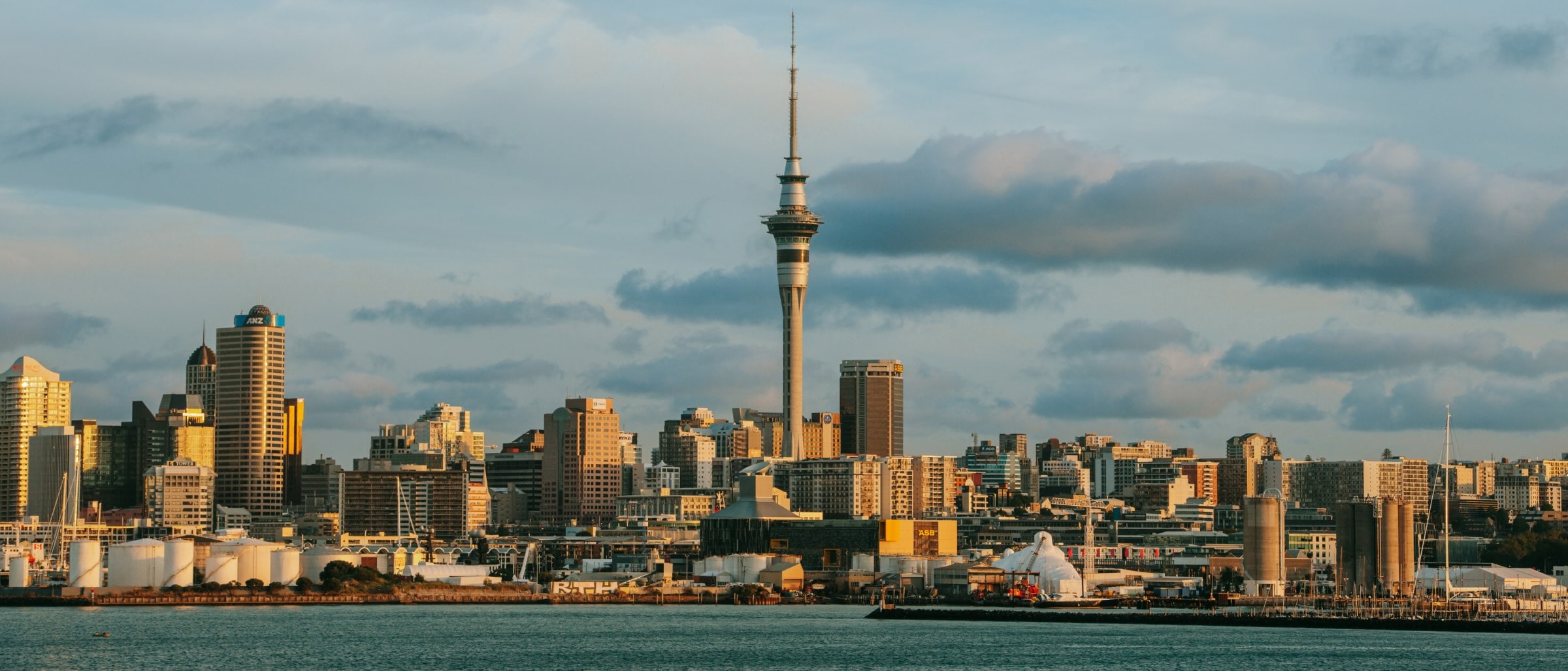 Skyline view of Auckland, New Zealand, featuring the Sky Tower rising above modern buildings near the waterfront under a partly cloudy sky.