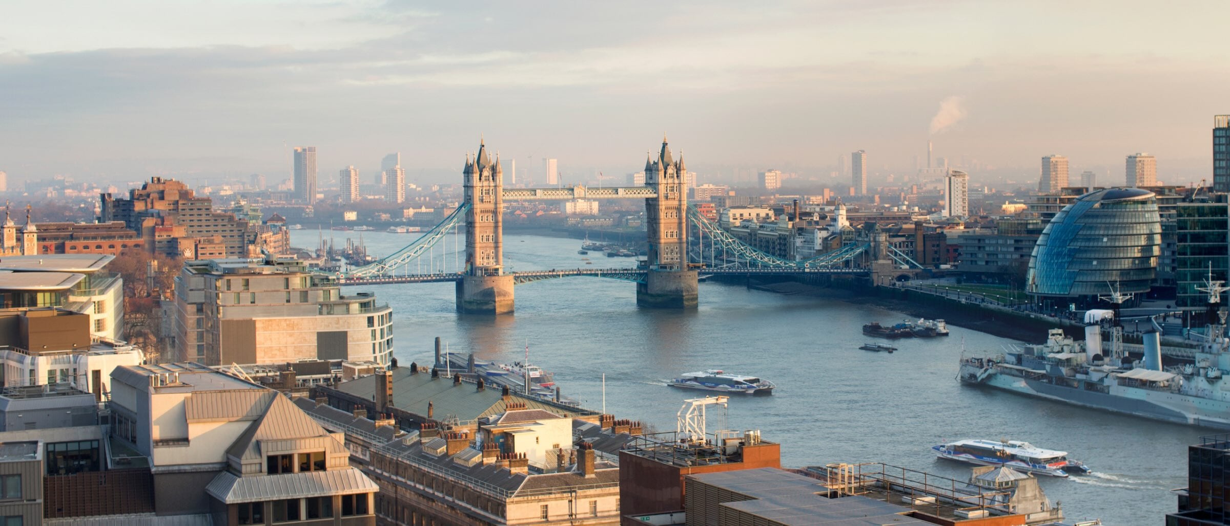 A wide view of London’s Tower Bridge spanning the River Thames, with modern buildings, boats on the water and a hazy city skyline in the background.