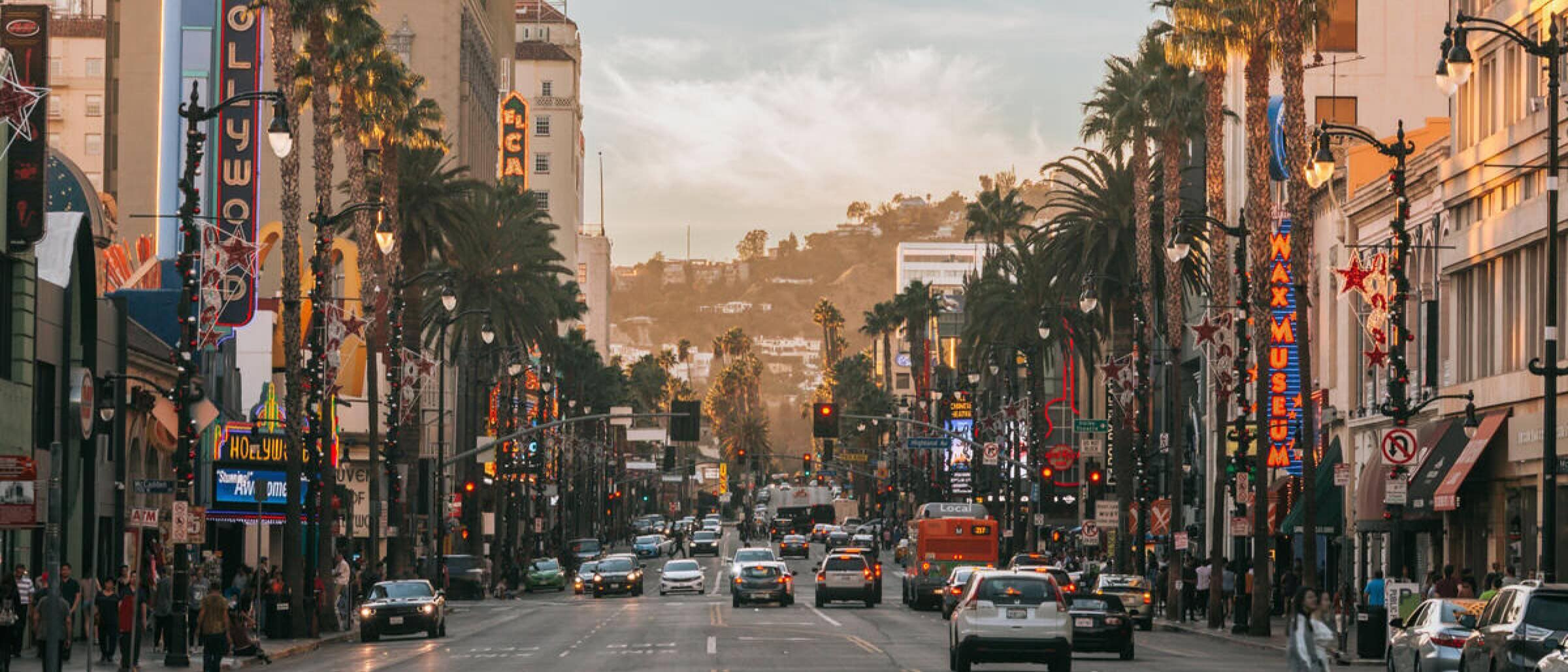 Busy street scene in Hollywood, Los Angeles, with cars, palm trees, neon signs, and the Hollywood Hills in the background at sunset.