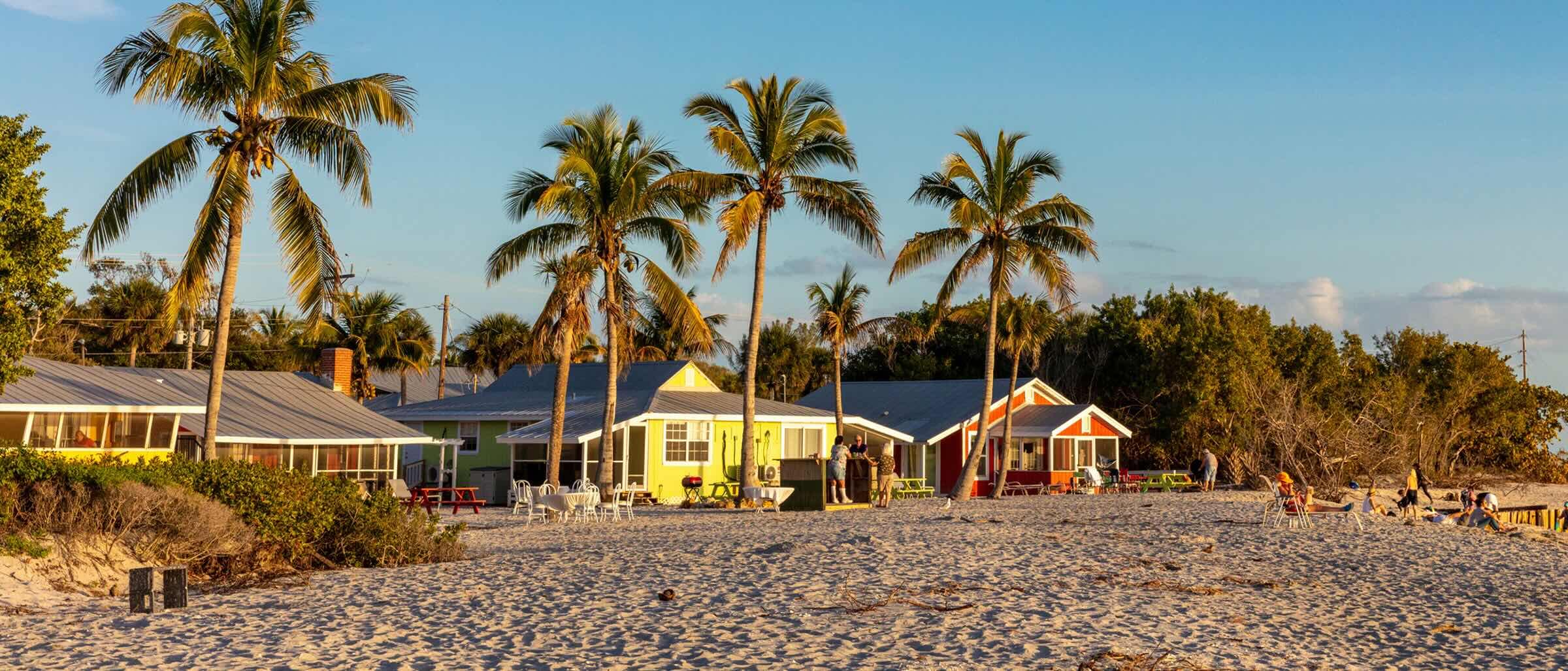 Colourful beach cottages with palm trees, set on a sandy beach at sunset. People are relaxing near the cottages and on the shore.