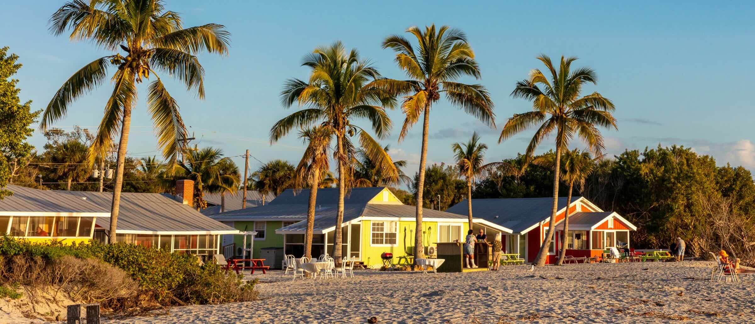 Colourful beach cottages with palm trees, set on a sandy beach at sunset. People are relaxing near the cottages and on the shore.