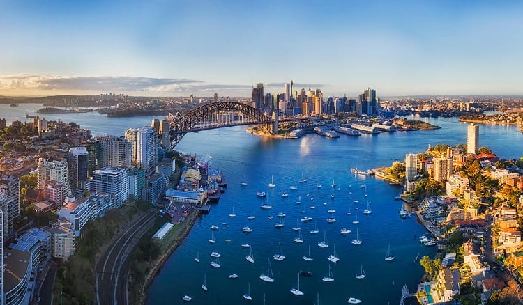 Coastal view of Sydney with the Harbour Bridge, Opera House and boats dotting the sparkling blue water on a sunny day.