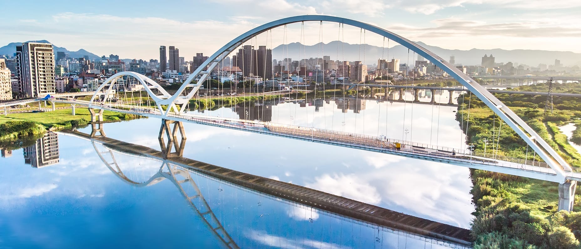 Modern arched bridge spanning a river in a cityscape with high-rise buildings and greenery reflected in the calm water.