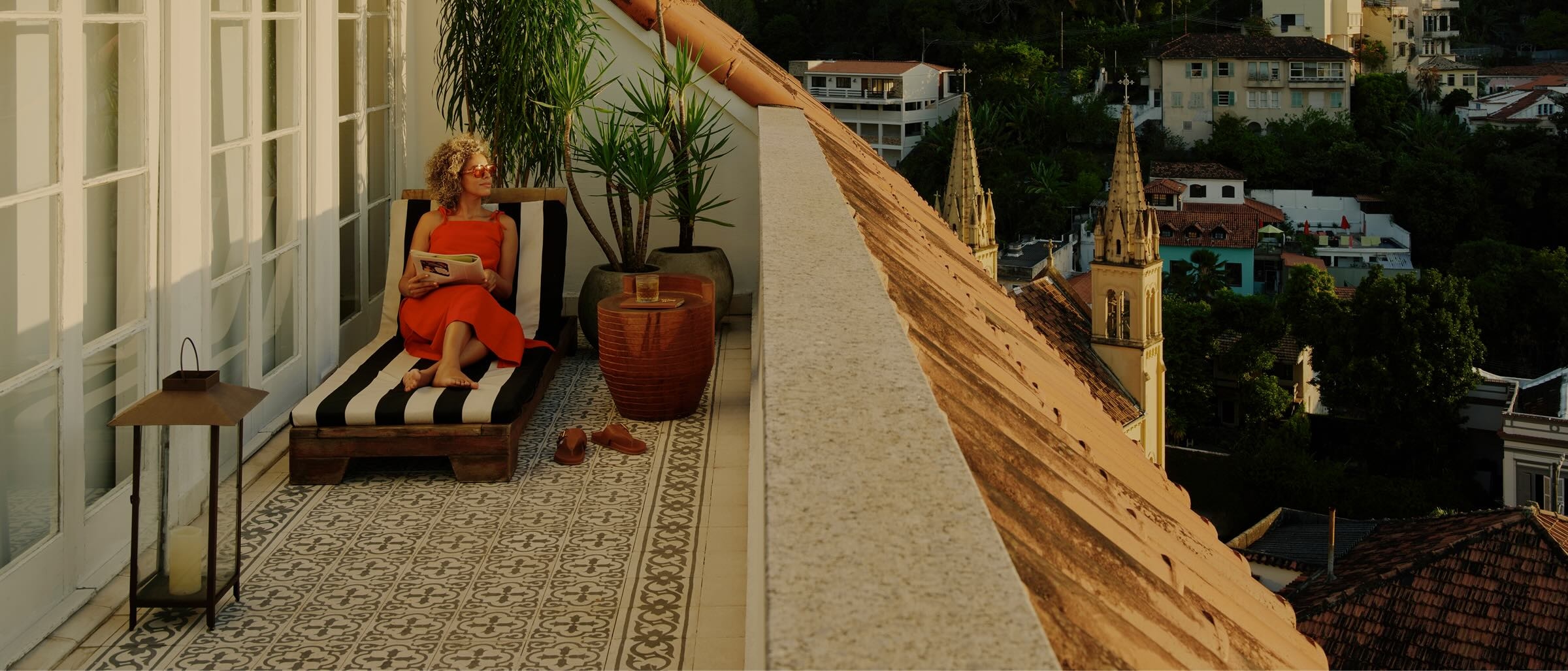 Uma pessoa relaxa com um livro no terraço privativo de um hotel com vista para uma cidade arborizada.