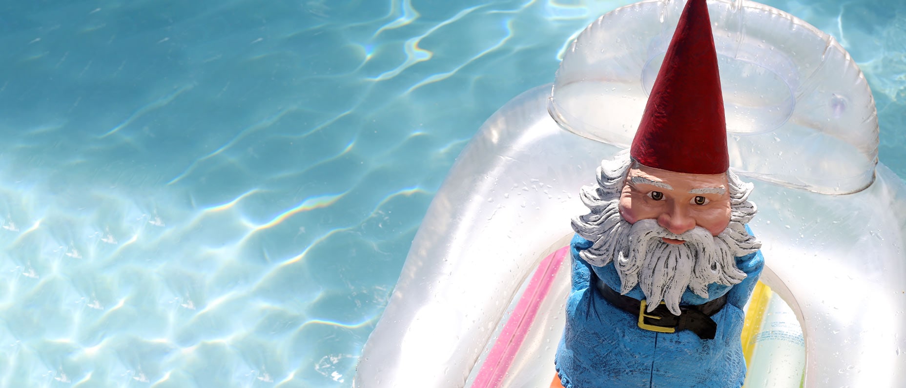 A figurine of a gnome with a red pointed hat, beard, and belted coat stands on an inflatable pool lounger floating on the water.
