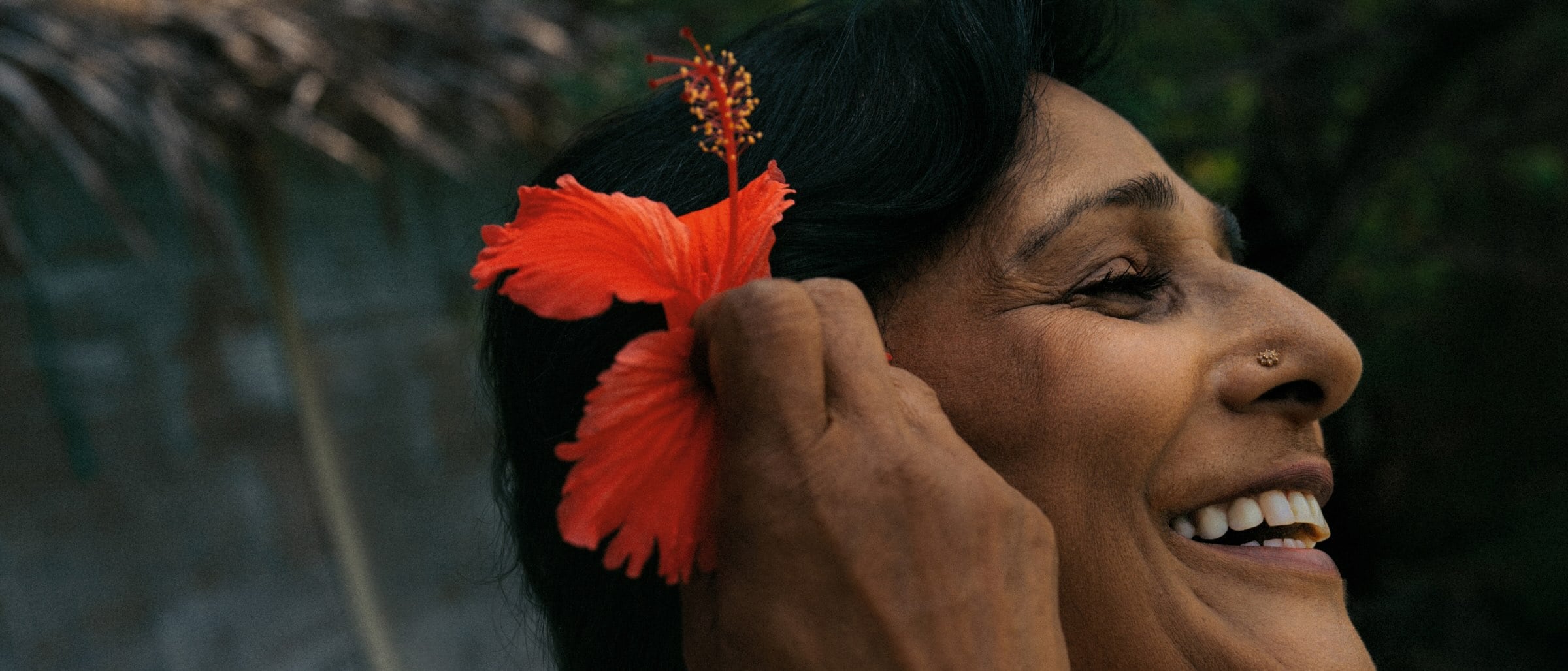Smiling person standing outdoors, holding a red flower in their hair.