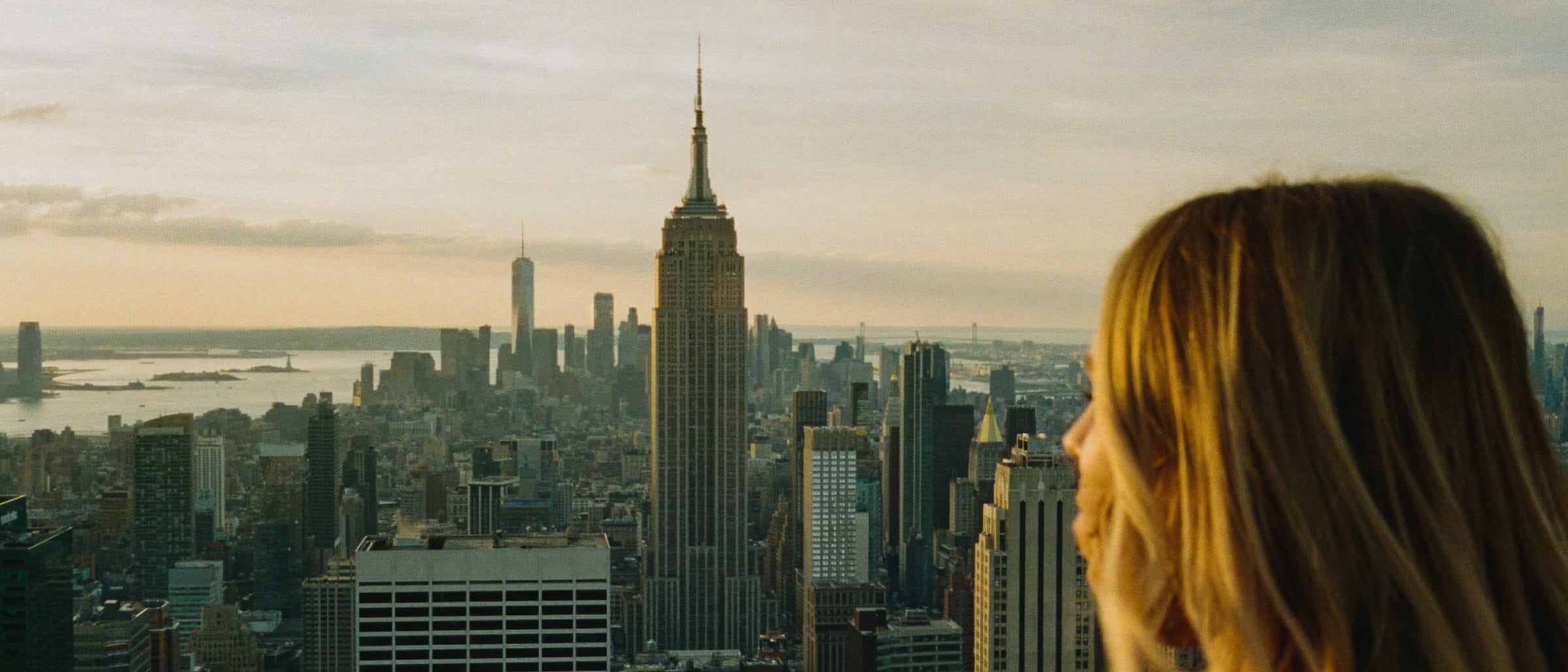 View of New York City skyline at sunset with a woman looking out from a rooftop.