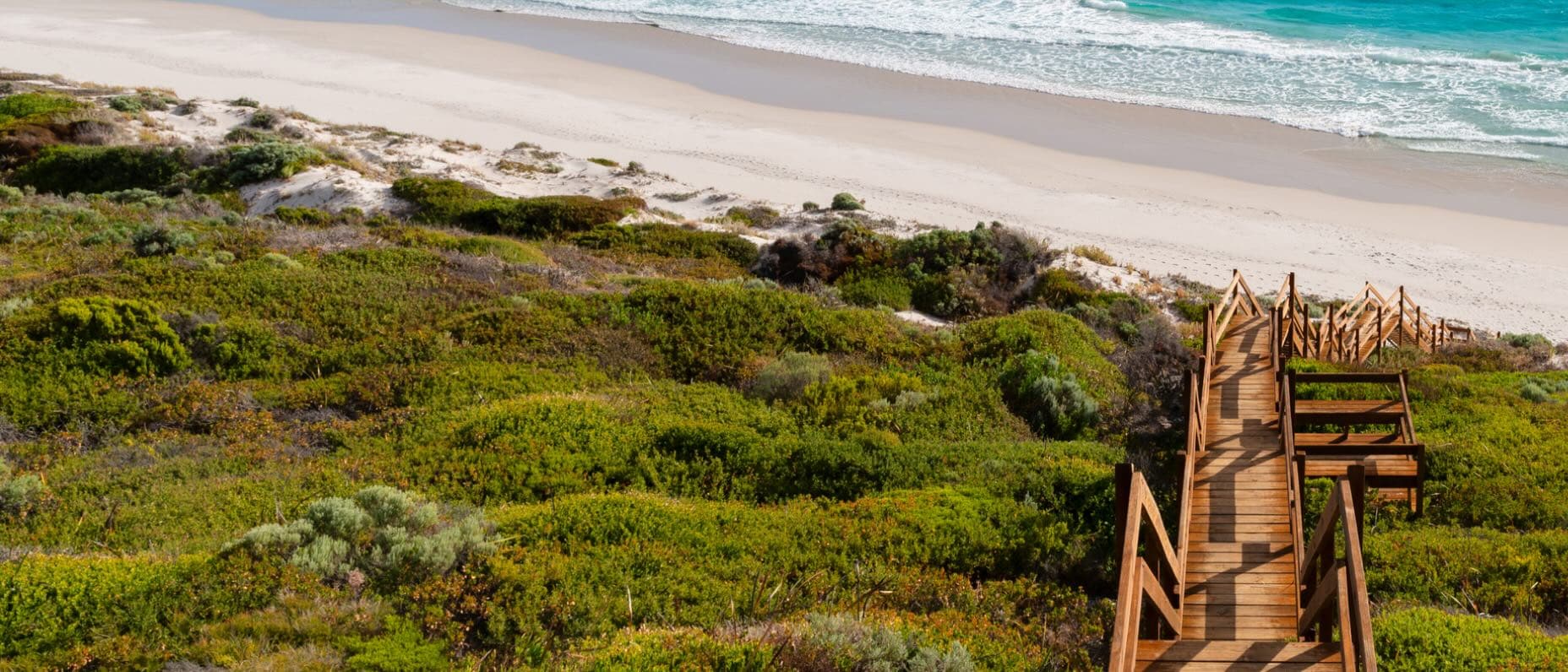 Wooden boardwalk leading through coastal greenery to a sandy beach with turquoise waves gently crashing onshore under a clear sky.