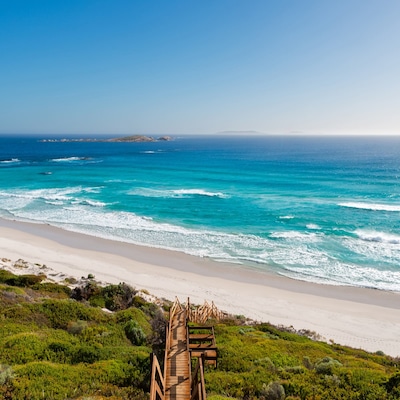 Two people walk on a sandy beach with surfboards, one blue and yellow, the other green and yellow. More people, buildings, and a clear sky are in the background.