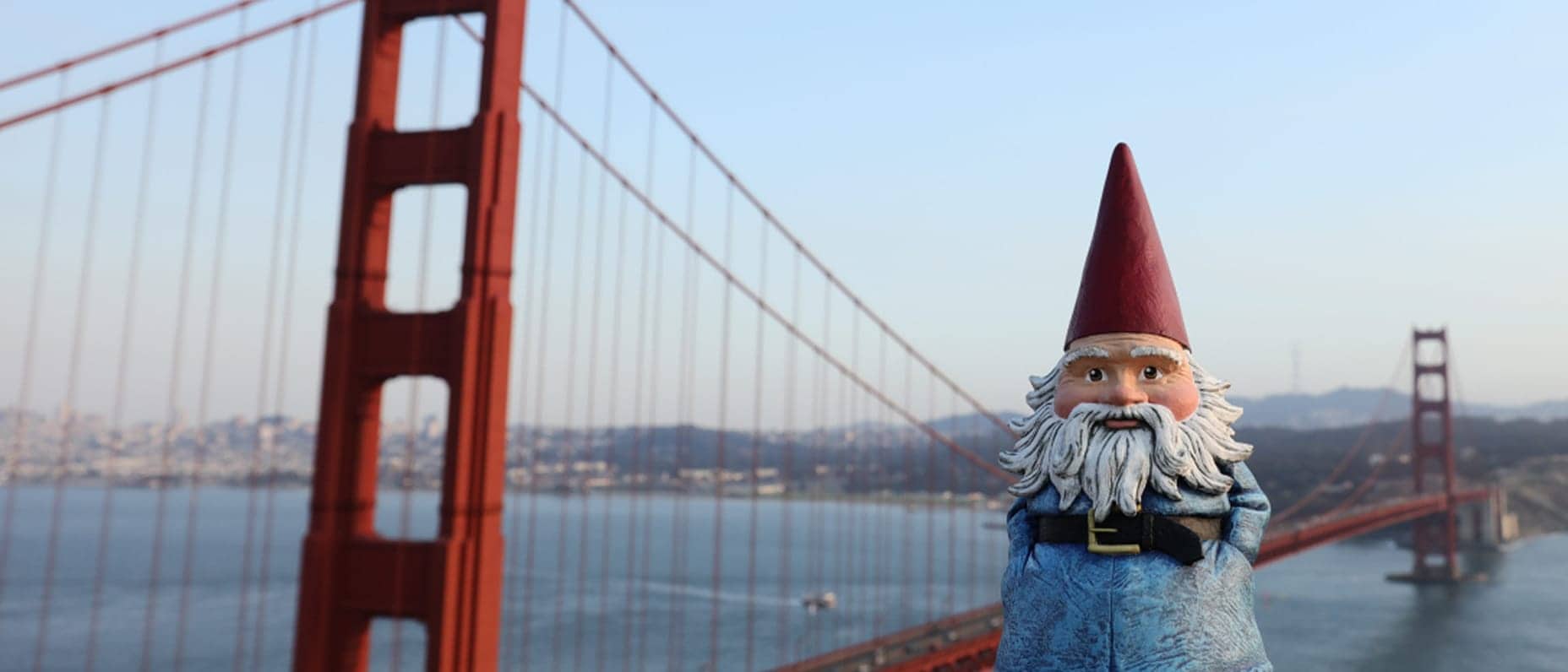 A gnome with a red pointed hat, white beard, and blue coat stands with arms crossed, while the Golden Gate Bridge and San Francisco Bay appear blurred in the background under a clear sky.