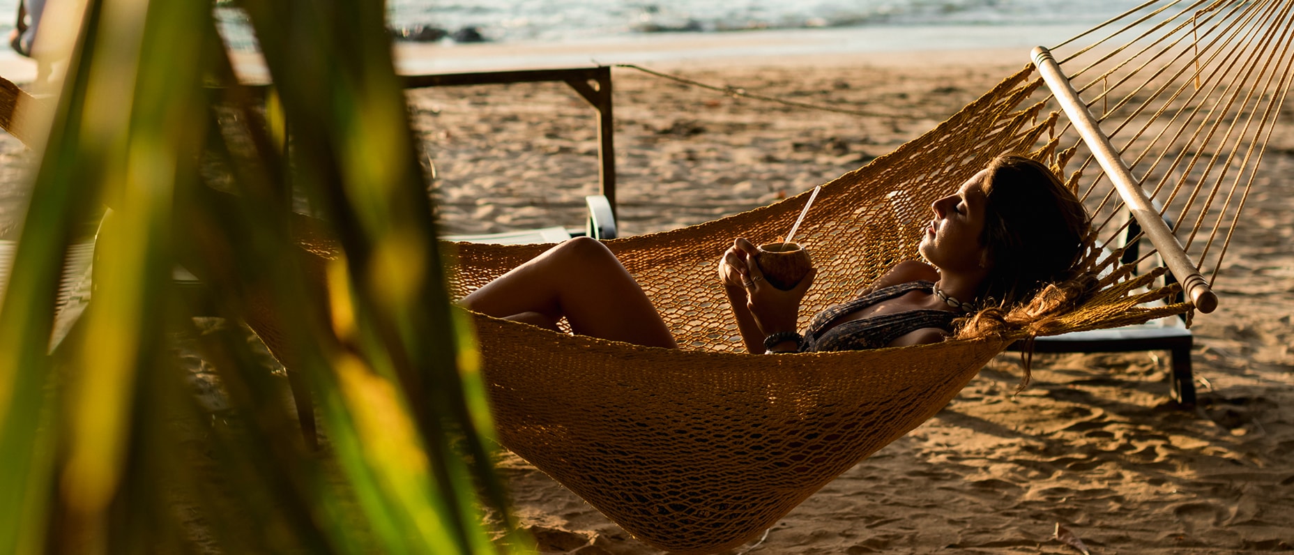 A woman relaxes in a hammock on a sunlit beach, drinking from a coconut. Palm leaves partially frame the scene, conveying a tropical, serene atmosphere.
