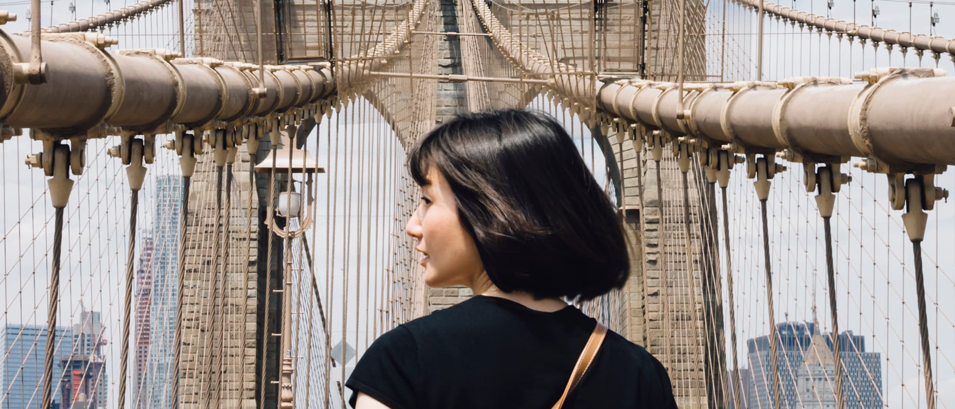 Une personne aux cheveux courts et foncés, vue de dos, se tient sur le pont de Brooklyn. Les câbles du pont l’encadrent sous un ciel clair, transmettant un sentiment d’aventure.