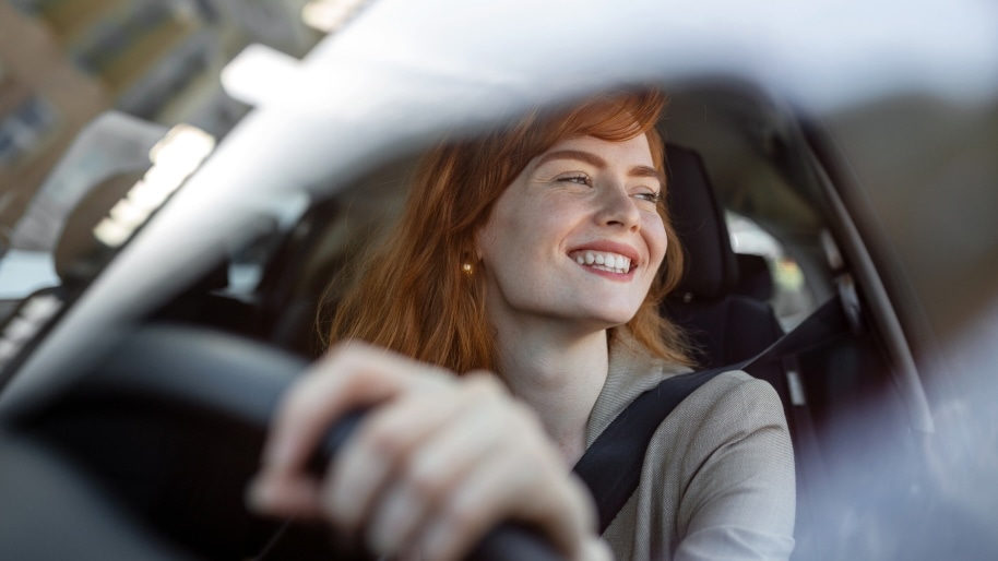 A woman with red hair drives a car, smiling joyfully. She grips the steering wheel, conveying a sense of freedom and happiness. The atmosphere is bright and uplifting.