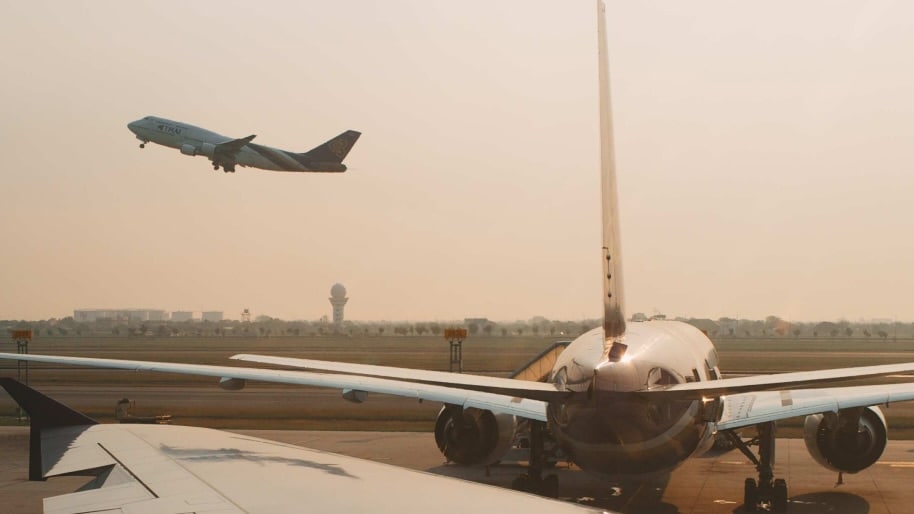 A plane taking off from a runway in the background, with another aircraft parked in the foreground. The scene is set under a warm, hazy sky.