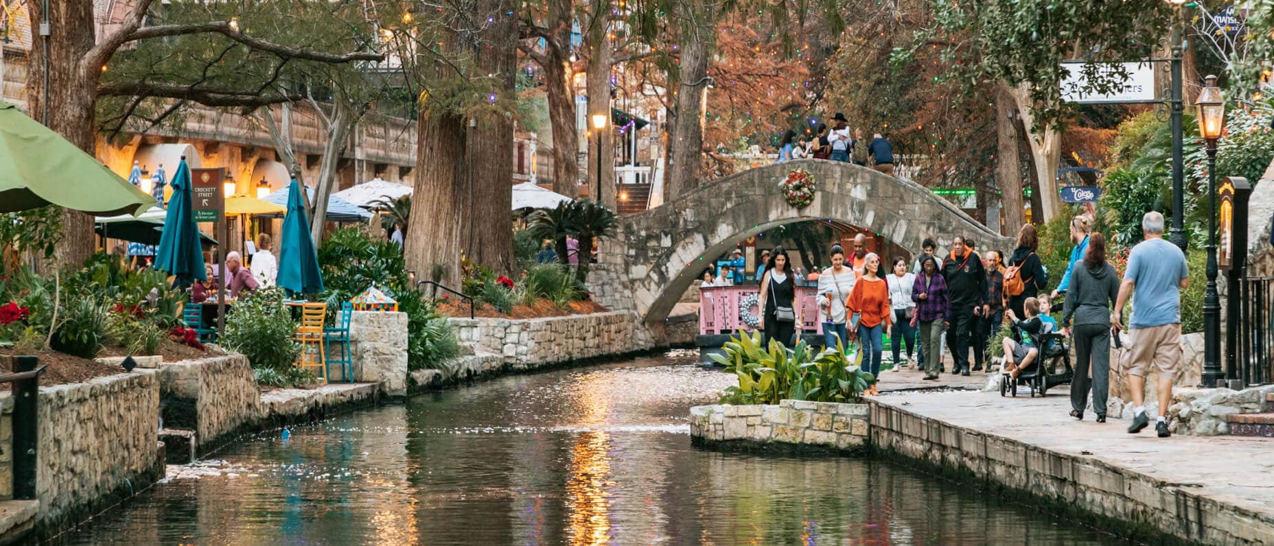 People strolling along the San Antonio River Walk surrounded by trees, outdoor cafes, and a stone bridge decorated with a wreath.
