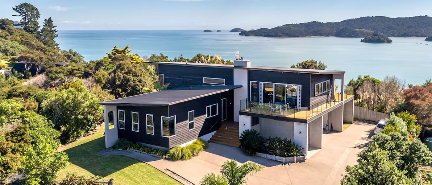 Modern hillside home with large windows and wraparound deck, surrounded by lush greenery and overlooking a calm bay dotted with small islands.
