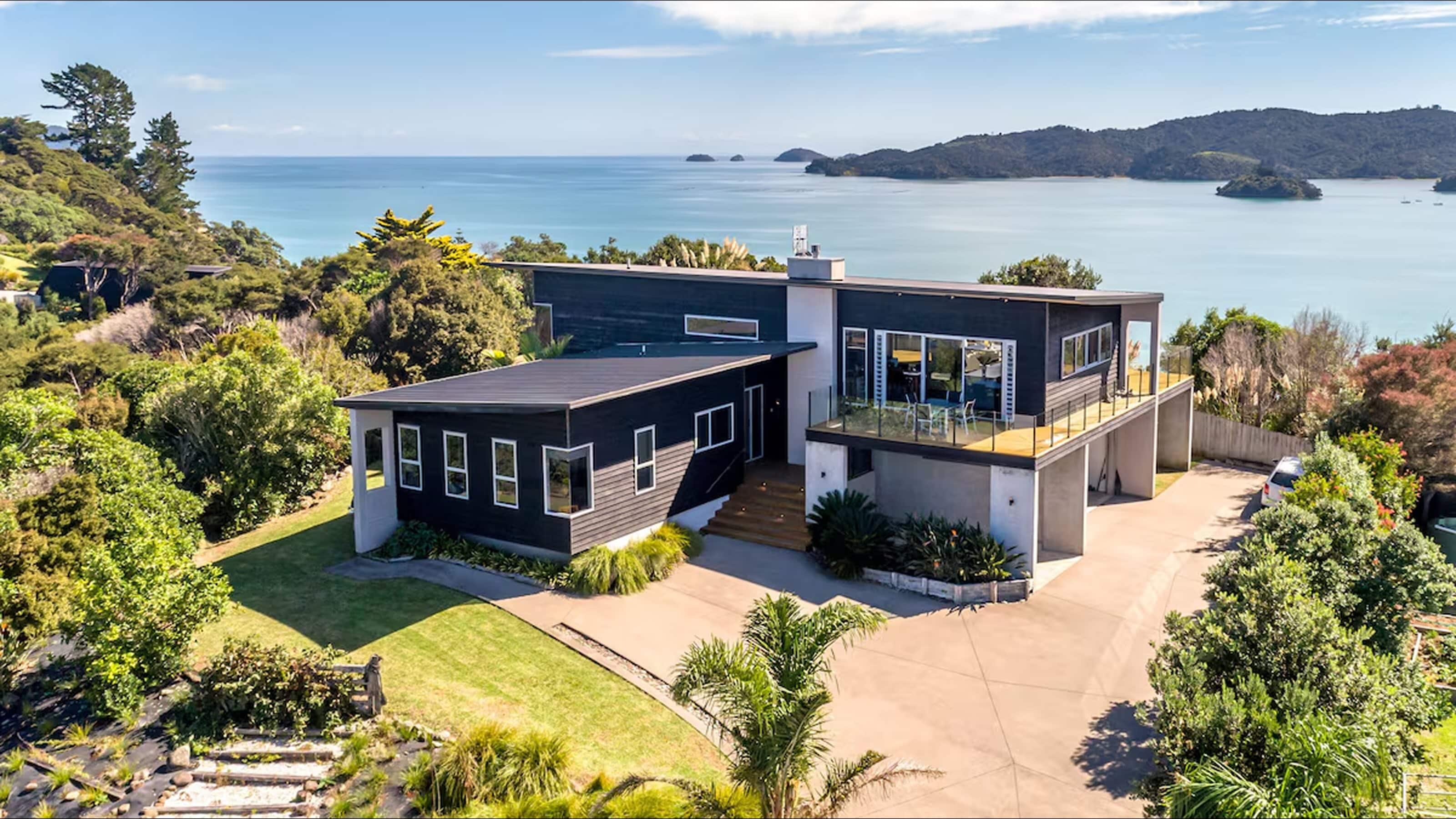 Sunny backyard with a rectangular pool, row of white loungers, outdoor dining set, and barbecue area beside a single-story house surrounded by trees.
