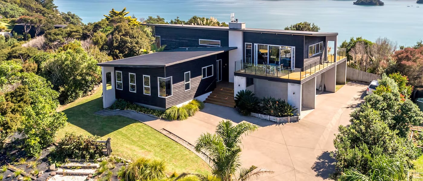 Sunny backyard with a rectangular pool, row of white loungers, outdoor dining set, and barbecue area beside a single-story house surrounded by trees.