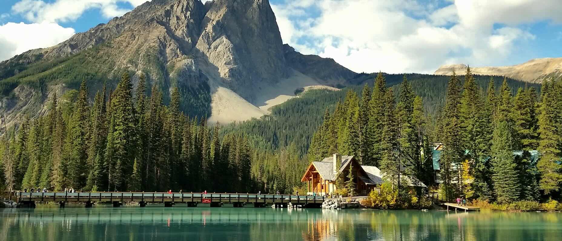 A group of visitors walk along a wooden footbridge, positioned above the turquoise waters of Emerald Lake in Yoto National Park. At the end of the footbridge, a log cabin is nestled amongst dense forest trees, while Mount Burgess rises dramatically in the background. 