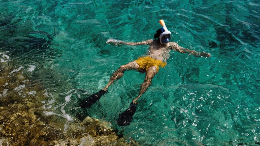 A man wearing a snorkel and yellow swim trunks lies on his back in turquoise sea water.