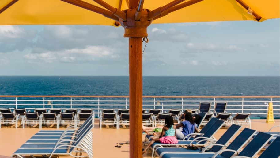 The pool deck of a cruise ship on a sunny day, with rows of navy blue sun loungers and a large yellow umbrella in the foreground. A family sits together on loungers looking out to sea, with a calm blue ocean and cloudy sky stretching to the horizon.