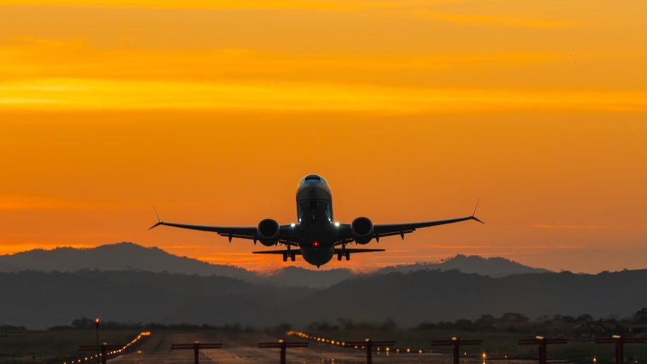 Sur une piste bordée de lumières, un avion décolle. Derrière l’avion, des collines sombres s’élèvent à l’arrière-plan.