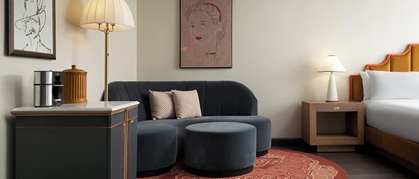 Quarto de hotel moderno com um sofá azul-marinho e um pufe ao lado da cama, luminárias de cabeceira, quadros emoldurados e uma decoração aconchegante.