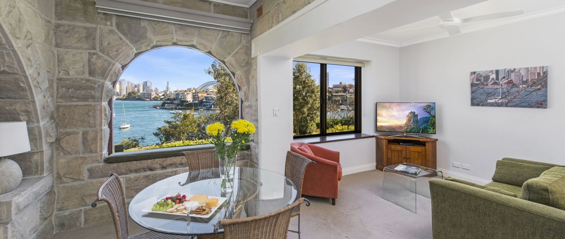 A living and dining space in a Sydney vacation rental, with stone walls in the dining area and a view from the window of Port Jackson and Sydney Harbour Bridge. 